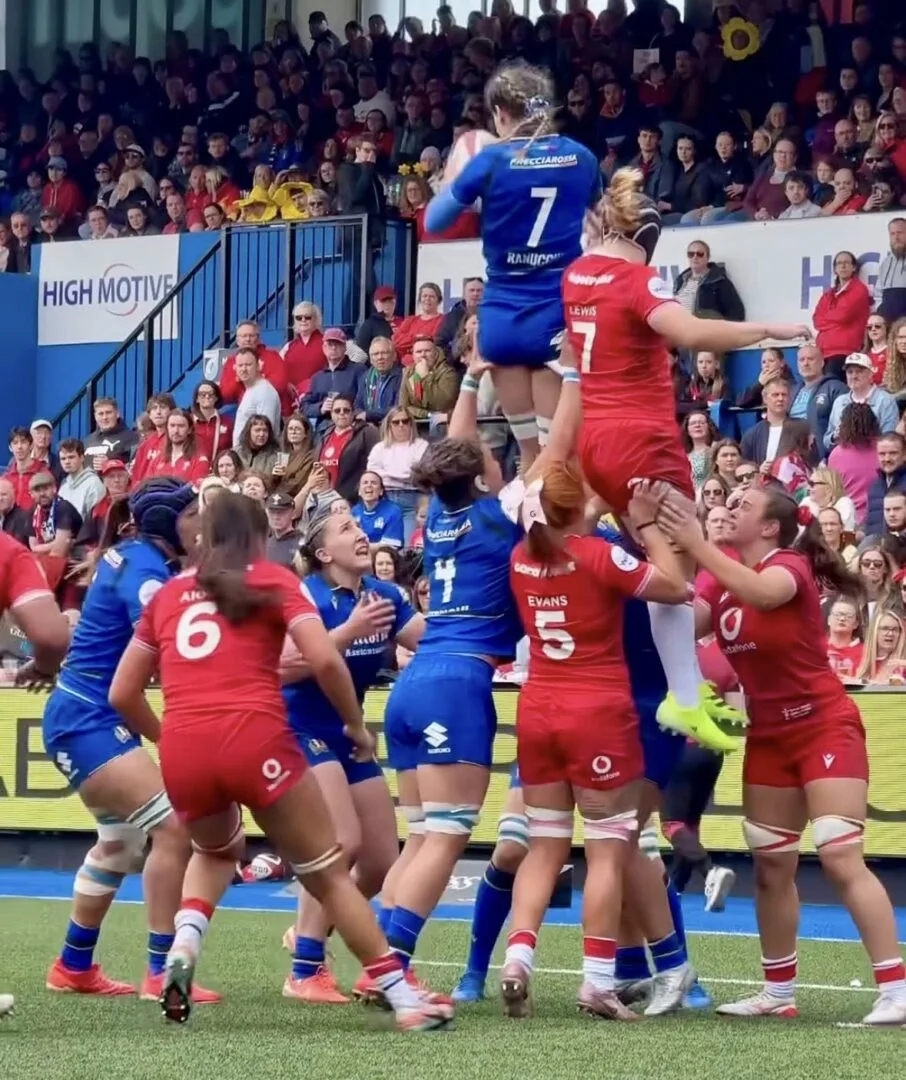 Giocatrici delle nazionali femminili di rugby di Italia e Galles saltano durante una touche mentre il pubblico assiste alla partita dagli spalti.