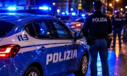 Agente della Polizia di Stato accanto a un'auto di servizio con lampeggianti blu accesi durante un intervento notturno in città. Sullo sfondo altre pattuglie e forze dell'ordine impegnate in un'operazione di controllo.