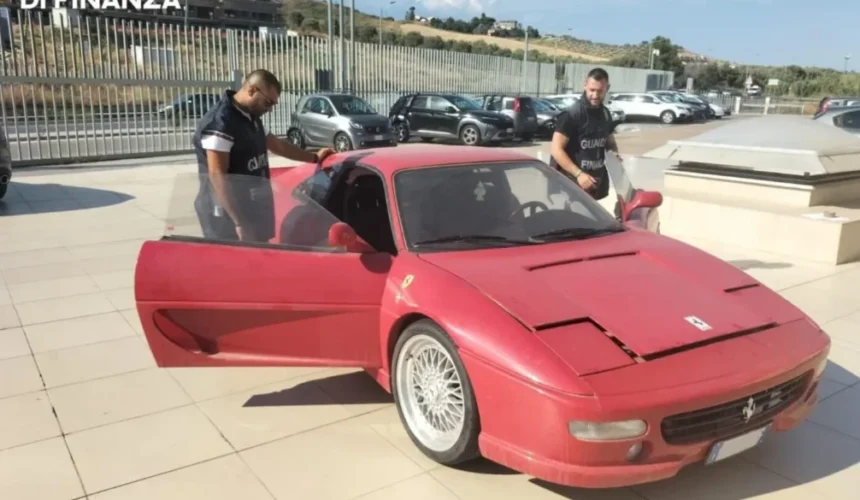 Due agenti della Guardia di Finanza controllano una Ferrari rossa parcheggiata in uno spazio aperto con altre auto sullo sfondo.
