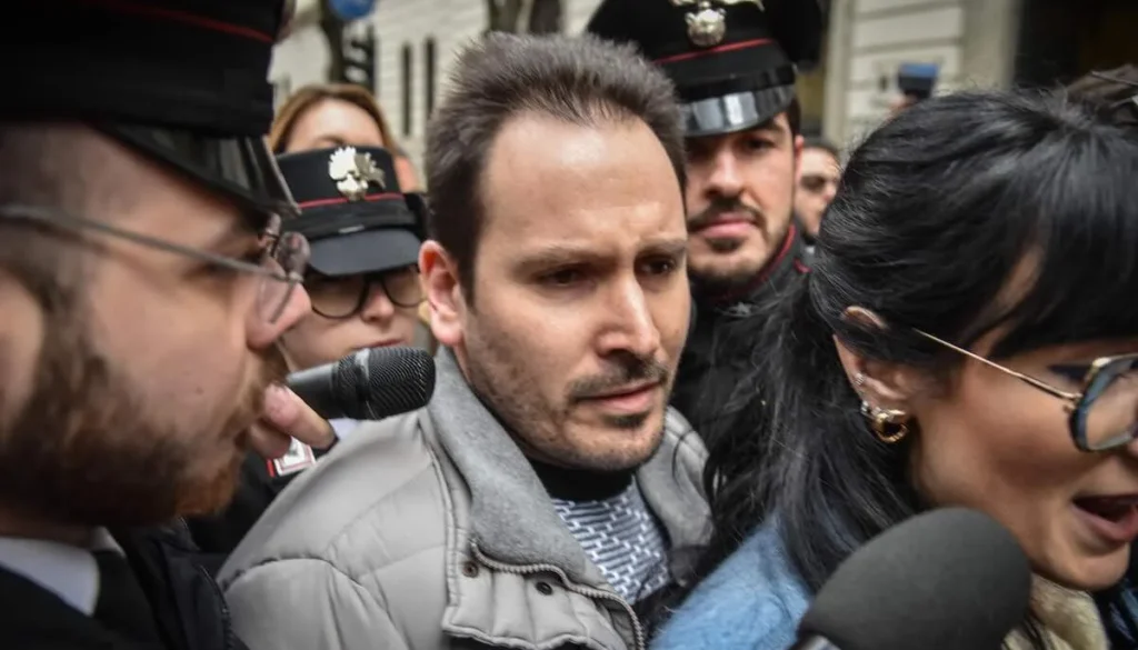 Uomo con giacca grigia circondato da giornalisti, microfoni e Carabinieri in una strada affollata durante un evento mediatico.