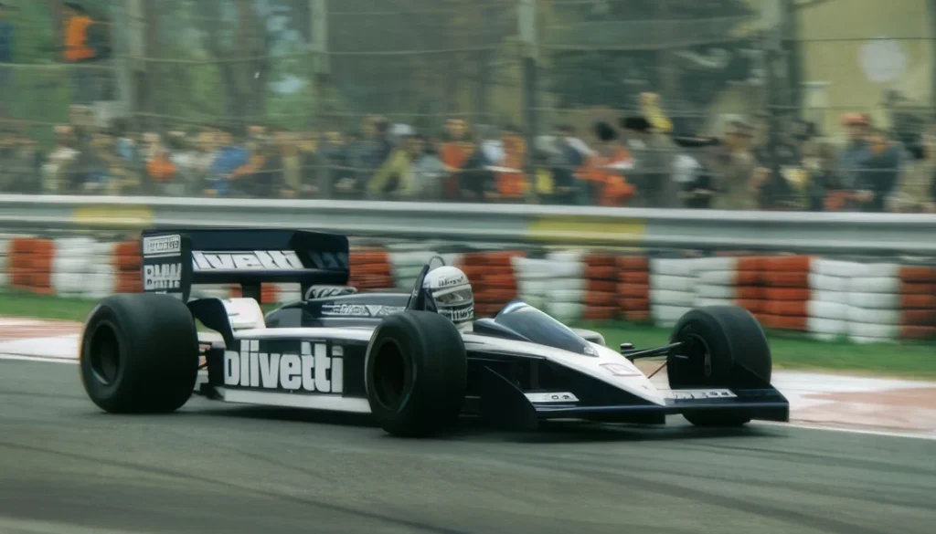 Auto storica di Formula 1 con livrea nera e bianca percorre una curva del circuito davanti agli spettatori dietro le barriere.