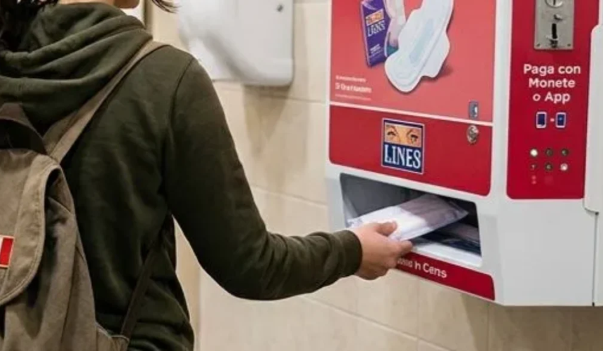 Persona con zaino mentre ritira un assorbente da un distributore automatico rosso installato in un bagno pubblico.