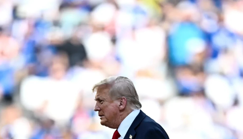 Donald Trump in abito scuro e cravatta rossa ripreso di profilo durante un evento pubblico con il pubblico sfocato sullo sfondo.