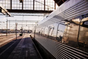 Treno moderno fermo in una grande stazione ferroviaria illuminata dalla luce calda del tramonto, con riflessi metallici sulle carrozze e binari che si estendono in prospettiva.