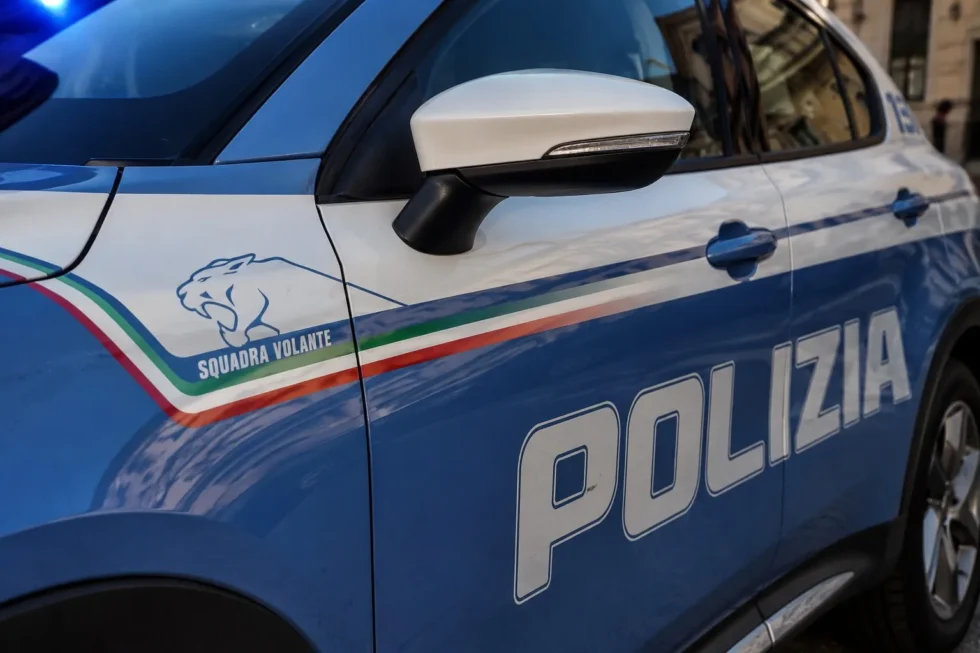 Primo piano di un'auto della Polizia di Stato italiana con la scritta "Polizia" ben visibile sulla fiancata, in un contesto urbano.
