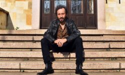 Uomo seduto sui gradini di un edificio con giacca in pelle nera e camicia fantasia, ritratto in un contesto urbano.