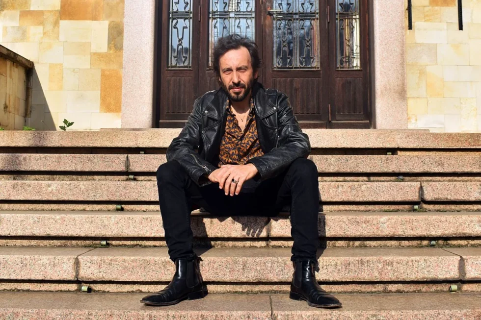 Uomo seduto sui gradini di un edificio con giacca in pelle nera e camicia fantasia, ritratto in un contesto urbano.